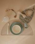 A flat lay of baby feeding items on a beige surface, featuring the mushie Silicone Baby Rattle Toy for a safe sensory experience.