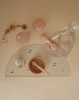 Flat lay of pastel baby items on beige: a bib, food-grade silicone feeding set with spoon and bowl, teapot, mushie Silicone Baby Rattle Toy, pacifier clip, soft block, and silicone finger toothbrush on a cherry mat for sensory play.