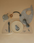A flat lay of baby feeding essentials on a light surface features a grey bib, bowl, sippy cup, spoon, food-grade silicone teether, pacifier clip with beads, and the mushie Silicone Baby Rattle Toy for safe sensory play.