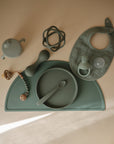 A flat lay of mushie's Silicone Baby Rattle Toy in sage green, paired with a matching plate, spoon, bib, sippy cup, teether, and beaded pacifier clip—all set on a coordinated mat against a neutral background for a calming sensory scene.
