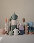 A pastel-toned display of mushie geometric toys—handled cups, textured balls, stackable arches—features the Silicone Baby Bottle Brush with silicone bristles for easy nipple cleaning, all arranged artistically on a neutral backdrop.