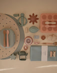 A collection of mushie Silicone Toddler Starter Spoons and pastel baby feeding essentials in soft blue, pink, and beige tones is neatly arranged on a beige background for independent feeding.