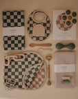 A flat lay of mushie Muslin Bibs in earthy tones, alongside a silicone teether, pacifier clip, utensils, teething rattle, swaddles, and checkered cloths, all neatly arranged on a beige background.