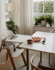 A bright dining area with a wooden high chair and a white table featuring the Mushie Silicone Fresh Food Feeder, a brown bowl, and a baby bottle. Large windows with sheer curtains and potted plants fill the space with sunlight.