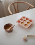 A bowl and mushie Silicone Toddler Starter Spoons, filled with yellow liquid from a pink silicone mold, sit on a white table. A baby toy and wooden chair complete this inviting setup for independent feeding.