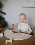 A smiling baby in a high chair sits at a wooden table with a bowl of snacks on the mushie Silicone Placemat, wearing a bib. A cup, spoon, and toy dog are nearby, with a green plant and framed map in the background.
