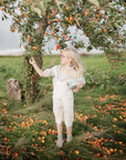 A young girl with long blonde hair and glasses stands under a fruit tree, reaching to pick fruit. She wears white overalls and holds a mushie Lunchbox with built-in compartments, while fallen fruit lies on the grass around her.
