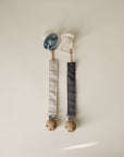The mushie Linen Pacifier Clip 2-Pack includes two pacifiers with fabric straps and wooden clips: one blue pacifier with a beige linen clip, and one white pacifier with a dark gray strap, both shown on a light background.