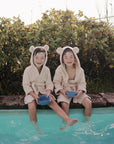 Two kids in bear ear robes by a pool hold mushie Square Dinnerware Bowls near green bushes.