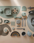 A flat lay of mushie silicone Toddler Starter Spoons, plates, bowls, bibs, teethers, stacking toys, and a rainbow in soft green, taupe, and gray arranged on a beige background.