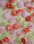 FRIGG Daisy Silicone Pacifier 2-Pack by FRIGG, shown in pink, green, and red, are displayed on a light surface adorned with small daisies and flower illustrations. Made in Denmark.