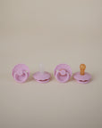 Two FRIGG Daisy Silicone Pacifier 2-Packs by FRIGG in pink and orange are lined up on a beige background. Each medical-grade silicone pacifier has a round handle, scalloped edges, and a baby comfort design.