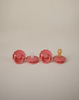 Four FRIGG Daisy Silicone Pacifiers from the FRIGG Daisy Silicone Pacifier 2-Pack, featuring medical-grade silicone nipples and daisy-shaped shields with pink bodies and multicolored knobs, are arranged in a row on a beige background.