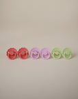 Four FRIGG Daisy Silicone Pacifiers by FRIGG, made from medical-grade silicone for comfort, are arranged in a row on a neutral background—two pink, one purple, and one light green—all with round handles and scalloped edges.