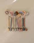 Six pastel-colored FRIGG Natural Rubber pacifiers with matching beaded clips are arranged on a beige surface. Each Denmark-made set features soft, muted tones and a round clasp. Brand: FRIGG. 6-pack included.