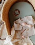 A beige knitted baby sweater, teether, pacifier clip, and teething ring are arranged on a green mattress inside a wicker bassinet with a soft cream cloth.