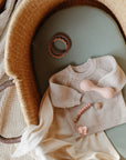 A beige knitted baby sweater, teether, pacifier clip, and teething ring are arranged on a green mattress inside a wicker bassinet with a soft cream cloth.