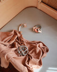 A light gray crib mattress with a tan blanket, a gray and peach silicone toy ball, a pacifier clip, and a teething ring with beads and animal shapes on top.