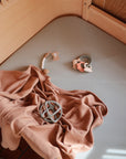A light gray crib mattress with a tan blanket, a gray and peach silicone toy ball, a pacifier clip, and a teething ring with beads and animal shapes on top.