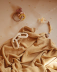 A soft beige blanket with textured dots is spread out, topped with a cream-colored teething toy. Nearby, a pacifier and a pastel beaded ring toy rest on a crib sheet decorated with small yellow stars.