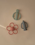 Three silicone baby teethers rest on a beige surface: a green robot, a red flower, and the mushie Rocket Rattle Teether in gray. Soft light casts gentle shadows, highlighting the playful design of each teething toy.