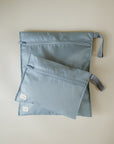 Two light blue mushie Water Resistant Wet Bags with small white tags and side straps rest on a light surface. The smaller pouch partially overlaps the larger, ideal for diaper bag storage.