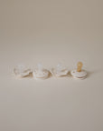 A set of four FRIGG Baby’s First Pacifier Moonlight Sailing (Cream) pacifiers, each with a silicone nipple, is arranged in a row on a plain light beige background.