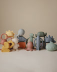 A collection of mushie silicone teethers, including the Rocket Rattle Teether, is arranged on a neutral background. The toys feature animal, flower, vehicle, and playful character designs in muted yellow, blue, green, pink, and orange.