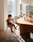 A young child sits in a high chair at a wooden dining table on the Mushie Splat Mat, eating breakfast as sunlight streams in, brightening the cozy room decorated with plants and candles.