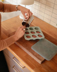 Someone pours liquid into the Mushie Baby Food Freezer Tray, a BPA-free silicone tray ideal for storing homemade baby food, set on a wooden cutting board in a kitchen with a tiled backsplash.