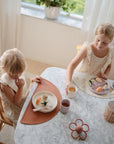 Two young children sit at a marble table set with food, drinks, and woven candle holders. Sunlight filters in through white curtains, and a mushie Silicone Suction Plate rests beside a potted plant on the windowsill.