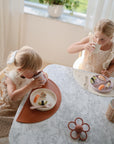 Two young children in light dresses sit at a marble table, enjoying sushi from plates placed on mushie Silicone Placemats. Sunlight shines through a window with white curtains, with a potted plant visible in the background.