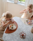 Two young children in light dresses enjoy a meal at a marble table with vegetables and bread, using the mushie Dinnerware Fork and Spoon Set. Natural light fills their cozy, tidy room as one drinks and the other holds her cup.