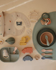 A selection of pastel baby feeding items and toys, featuring mushie Silicone Placemats, bowls, spoons, an animal-print bib, teething rings, and toy vehicles—all arranged neatly on a beige surface.