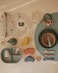 A flat lay of pastel-colored mushie Silicone Toddler Starter Spoons and baby feeding essentials—bibs, bowls, teether rings, pacifier, sectioned plates, and silicone toy vehicles—designed to encourage independent feeding.