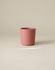 A matte pink mushie Dinnerware Cup for kids rests on a light beige surface with a neutral background, softly lit by natural light—safe and non-toxic for everyday sipping.