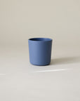 A minimalist photo of a plain blue mushie Dinnerware Cup, non-toxic, sits on a light gray surface with a neutral background as soft light falls across it.