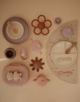 A flat lay of mushie pastel-colored silicone Lunchboxes and matching feeding items, including plates with compartments, bowls, cups, spoons, a bib, teether, and placemat, all neatly arranged on a beige background.