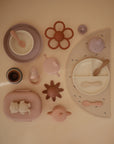 A flat lay of pastel silicone baby feeding items—including plates, bowls, spoons, the Mushie Trainer Sippy Cup with easy-grip handles, a flower teether, bib, and placemat—arranged neatly on a beige background.