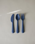 The mushie Dinnerware Cutlery Set, featuring a blue knife, spoon, and fork made from non-toxic, dishwasher-safe plastic, is arranged side by side on a light grey surface under a diagonal streak of light.