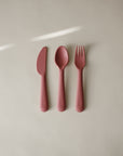 The mushie Dinnerware Cutlery Set, featuring a pink knife, spoon, and fork, is laid out on a light beige surface with sunlight above. Each non-toxic piece is dishwasher-safe for daily use.