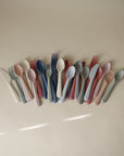 The mushie Dinnerware Fork and Spoon Set, featuring colorful, non-toxic, dishwasher-safe plastic utensils in pastel pink, blue, green, and white, is arranged in a curved line on a beige surface.