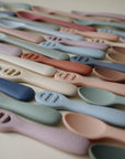 Pastel-colored Mushie First Feeding Baby Spoons are arranged in rows on a light surface. Each silicone spoon has a rounded handle embossed with the brand name, ideal for self-feeding and teething babies.