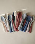 The mushie Dinnerware Fork and Spoon Set features neatly arranged pastel pink, blue, and beige utensils made from non-toxic plastic, displayed on a neutral surface.