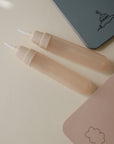 Two pale pink silicone baby utensils with white tips sit on a light surface, next to pastel notebooks and sustainable art supplies, including the mushie Reusable Water Color Pad.