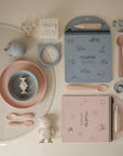 A flat lay of pastel-colored baby items, featuring the mushie Reusable Water Color Pad with a refillable paintbrush, bowls, spoons, teethers, a sippy cup, gift box, coloring pad with pen, and bib—neatly arranged on a light background.