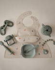 A flat lay of mushie's Silicone Placemat, silicone bib with dinosaur print, bowl and spoon, sippy cup, teether, and pastel baby accessories arranged on a neutral background.