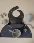 A flat lay of blue-grey baby feeding items, including a colorful dot bib, silicone bowl and spoon, sippy cup, pacifier clip, teether, two finger toothbrushes on mushie Silicone Placemats.