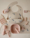 A flat lay of pastel pink and cream baby feeding and teething items, including a floral bib, bowl with spoon, silicone teether, pacifier, training cups, and phone-shaped toy—all arranged on a mushie Silicone Placemat.