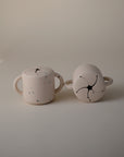 The mushie Snack Cup includes two cream-colored ceramic mugs with double handles and subtle colored speckles; one mug displays a black flower-like design. These are the ideal size for toddlers moving on from a silicone snack cup.